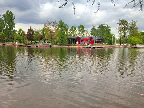 Serene lakeside park scene featuring a calm body of water, wooden docks, and a bright red modern structure among green trees. Overcast skies soften the light, creating a peaceful, reflective atmosphere suitable for outdoor, leisure, and lifestyle stock use.