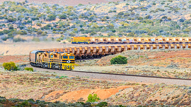 Pimba, Australia - September 4, 2023:  Telephoto view of an empty Aurizon Wirrida iron ore train shimmering in the heat haze as it snakes through the curves of Wirrappa in South Australia's remote north. Two modern powerful diesel locomotives designed for the harsh conditions, lead the train with a third attached to the rear. Behind the locomotives are in-line fuel tanks so that the locomotives can be fuelled on the move. Behind the fuel tank is a specially converted passenger car to with accommodation facilities for a second train crew to allow relay working between the crews across the vast distances of Australia.