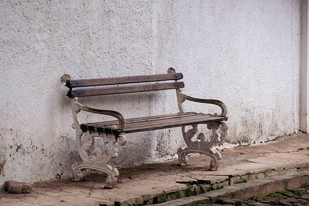 An empty vintage-style park bench with dark wooden slats and ornate white wrought-iron legs sits on a cracked stone walkway. The bench is positioned directly against a textured, weathered white stone wall.