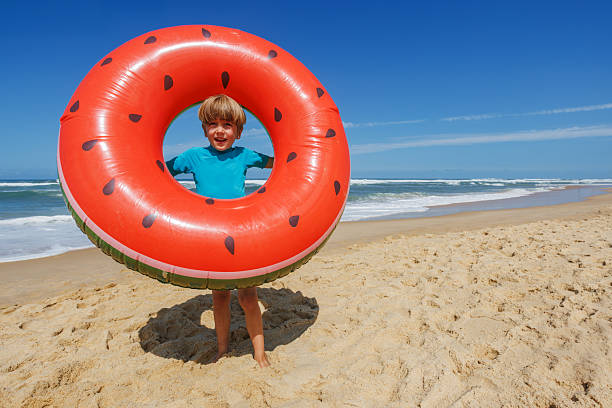 A cheerful child stands on the sandy beach holding a large watermelon-shaped float, enjoying a sunny day by the ocean waves, the clear blue sky filled with excitement and joy