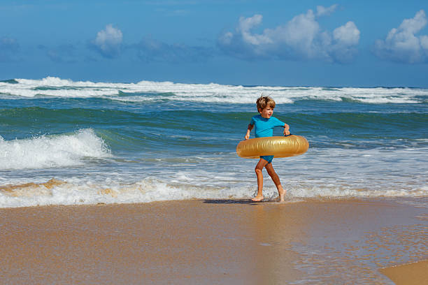 A young child joyfully walks along the sandy beach holding a golden inflatable ring while waves crash gently in the background under blue sky