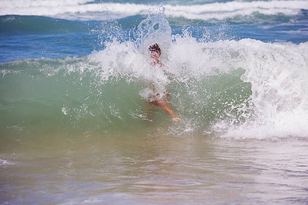 A teenage boy is laughing and shouting at the camera as a huge wave engulfs him on a beach in Normandy, France. Shot with Canon R5 on a sunny summer day.