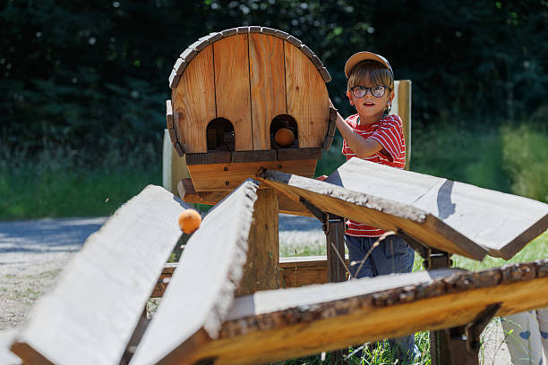 A young child engages with a wooden toy marble run designed for outdoor play, enjoying a sunny day in a natural setting