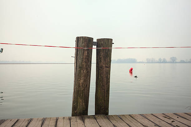 Mooring poles with a striped tape on them, and buoys floating on a still lake casted in the water, framed by the poles, on a cloudy and misty day
