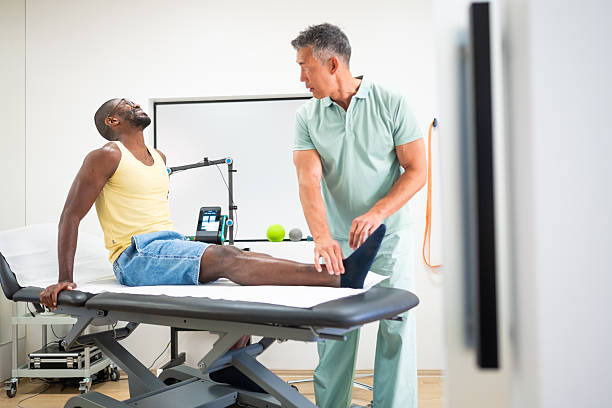 Asian therapist attentively checking ankle of mid adult Black male patient in clinic setting as patient sits on examination table.