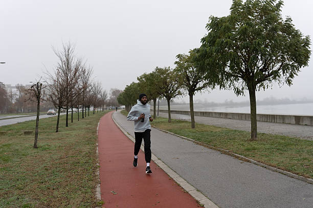 Young athletic man jogging outdoors along a tree-lined riverside path, wearing hoodie and beanie. Urban running workout, endurance training, healthy active lifestyle in the city