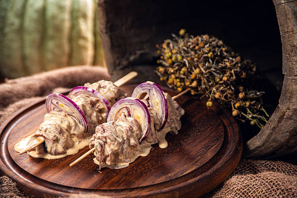 Close-up image of raw marinated beef skewers layered with red onion slices, arranged on a rustic wooden serving board. The beef is coated in a creamy herb marinade, creating a rich texture and artisanal appearance. Natural light and shallow depth of field enhance the gourmet food styling.Perfect for concepts such as kebab preparation, barbecue, grilling, meat marination, home cooking, traditional cuisine, butcher shop advertising, and restaurant menus. Ideal for commercial food photography, recipe blogs, culinary editorials, and premium meat branding.