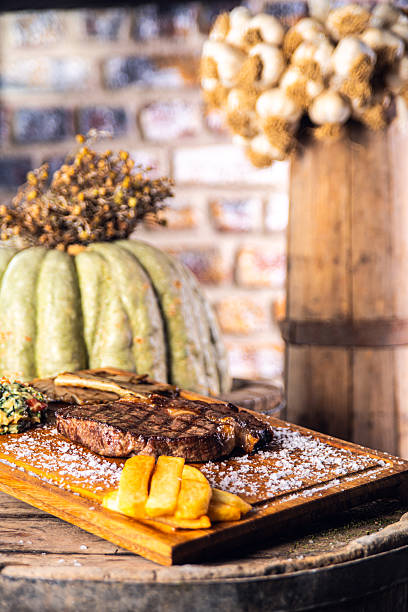 High-quality food photography featuring a perfectly grilled ribeye steak served on a rustic wooden board, garnished with crispy golden potatoes, coarse sea salt, and gourmet side vegetables. The scene is styled with a warm, rustic steakhouse atmosphere, brick wall background, and natural props that emphasize premium meat quality and artisanal presentation.Ideal for restaurant menus, steakhouse branding, gourmet food advertising, hospitality marketing, culinary blogs, and luxury dining promotions. The shallow depth of field and warm lighting highlight the juicy texture, grill marks, and upscale dining experience.