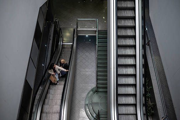 Two women on an escalator, one holding a shopping bag, moving down in a contemporary urban shopping center