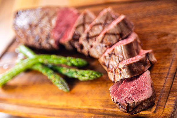 Professional food photography of sliced medium rare beef steak served on a wooden cutting board, accompanied by fresh green asparagus. The image features a shallow depth of field, highlighting the tender pink interior, seared exterior, and juicy texture of the steak, while the background remains softly blurred.Warm lighting and rustic styling create an appetizing and premium look, ideal for steakhouse menus, fine dining promotions, gourmet food advertising, restaurant branding, culinary blogs, recipe covers, and meat or protein-focused marketing content. The composition conveys quality beef, professional cooking, and modern gastronomy.