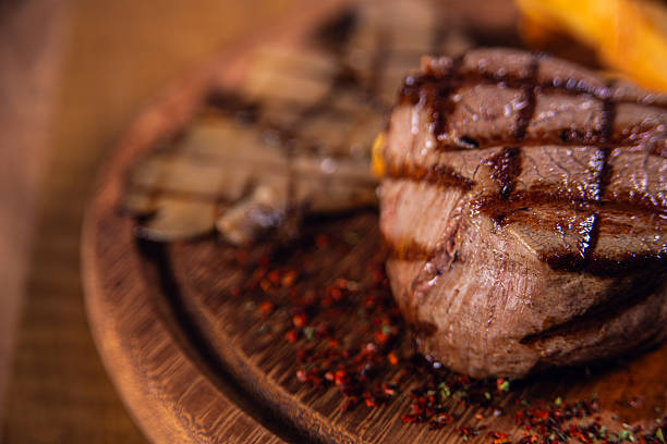 Close-up gourmet food photography of a grilled beef steak with pronounced grill marks, served on a round wooden plate. The steak appears juicy, tender, and perfectly seared, with visible texture and rich brown tones that emphasize premium quality meat and professional grilling technique.The shallow depth of field and warm lighting create a luxurious, appetizing atmosphere, ideal for steakhouse branding, restaurant menus, food advertising, culinary blogs, and commercial stock photography. The rustic wooden surface and subtle seasoning details enhance the authentic, high-end dining experience.Perfect for use in restaurant promotions, menu design, food delivery apps, social media marketing, cooking concepts, and premium food-related visual content.
