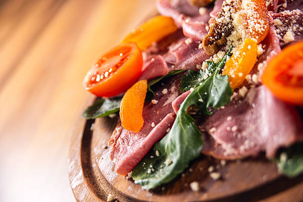 Artistic close-up food photography of a refined beef carpaccio dish presented on a rustic wooden board. Thinly sliced raw beef is delicately layered with fresh arugula leaves, halved cherry tomatoes, grated cheese, and vibrant citrus segments, creating an elegant balance of colors and textures.The shallow depth of field and warm, soft lighting highlight the freshness and tenderness of the beef while drawing attention to the natural ingredients and gourmet plating. The angled composition adds a sense of sophistication and movement, making the image visually engaging and appetizing.Ideal for concepts related to Italian cuisine, Mediterranean food, gourmet appetizers, raw beef dishes, fine dining, healthy eating, restaurant menus, food advertising, culinary blogs, and premium stock photography. Suitable for commercial use, editorial design, branding, and high-end food marketing projects.