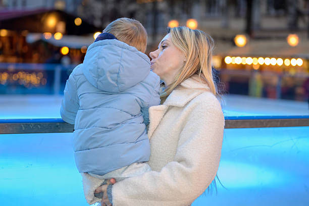 Mother holding and kissing her child outdoors during a winter evening with festive lights and blurred Christmas market background.