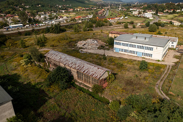 Aerial view of a rusted, abandoned warehouse next to a modern industrial building, set in a grassy, semi-rural area with scattered vegetation. The juxtaposition of decay and development is framed by distant hills and a quiet residential town in the background.