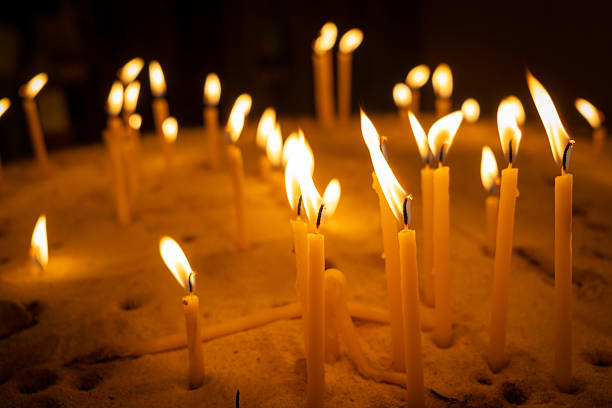 A group of burning candles stands in a bed of sand, their flickering flames casting a warm, golden glow. The wax drips down as the firelight creates a peaceful and spiritual atmosphere, symbolizing reflection and devotion