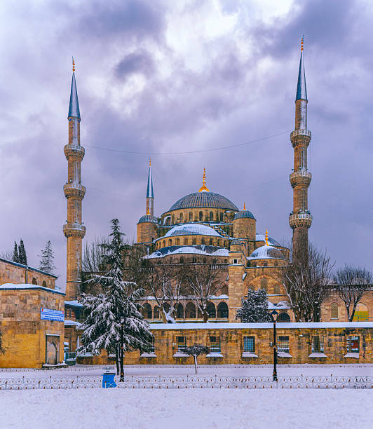 The Blue Mosque (Sultan Ahmed Mosque) covered in fresh snow in Istanbul, Turkey. A peaceful winter cityscape with a lone person walking in the foreground, highlighting historic Islamic architecture under a soft overcast sky. The person is not identifiable.