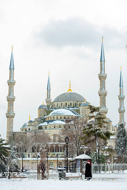 The Blue Mosque (Sultan Ahmed Mosque) covered in fresh snow in Istanbul, Turkey. A peaceful winter cityscape with a lone person walking in the foreground, highlighting historic Islamic architecture under a soft overcast sky. The person is not identifiable.