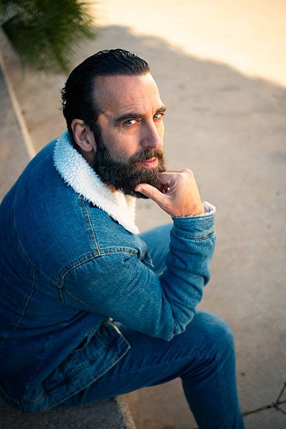 Man with beard wearing denim jacket thinking while sitting outdoors