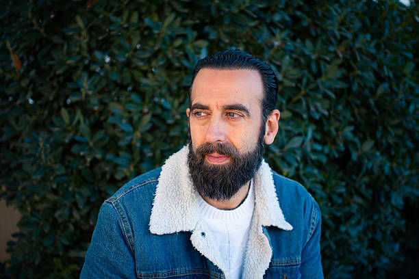 Man with beard looking away in a denim jacket with green foliage background