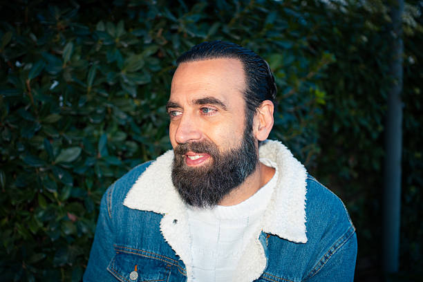 Male with beard and denim jacket looking away, standing against green foliage outdoors illuminated with direct flash