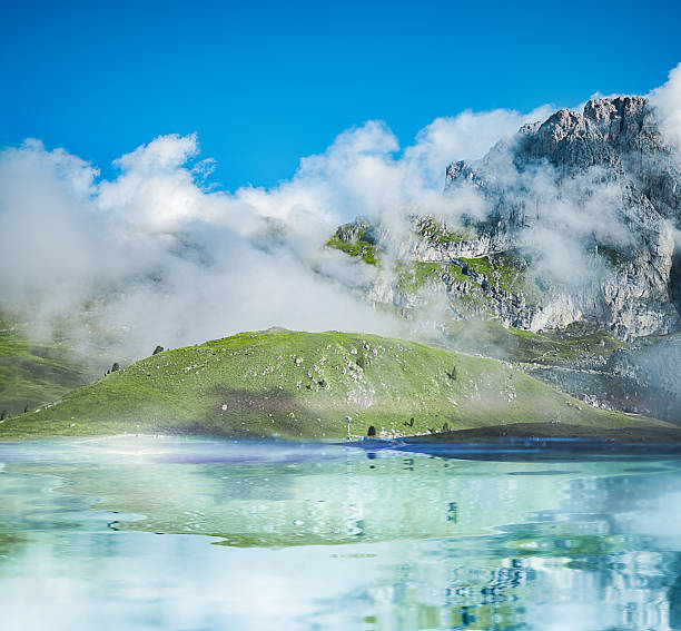 Scenic alpine landscape with a calm turquoise lake reflecting green grassy hills, dramatic rocky mountain peaks and drifting low clouds in bright summer weather, with wide blue sky and copy space.