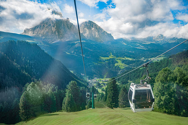 Scenic summer view from a gondola cable car descending over a green alpine valley with dense pine forests, small villages and rugged mountain peaks under dramatic clouds and blue sky, popular travel and tourism destination.