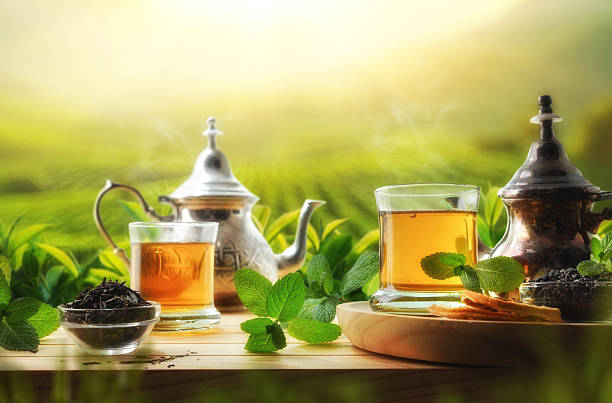 Glass cups of hot green tea on a wooden table with dried tea leaves and mint leaves and several Moroccan-style metal teapots and a sunny outdoor background with a tea plantation. Front view.