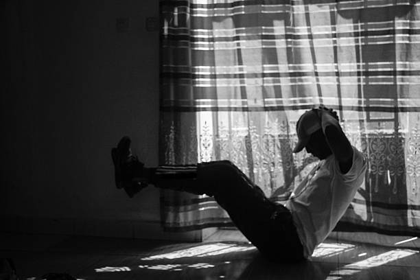 A black and white silhouette of a man balancing in a seated core position with hands behind his head. Backlit by natural sunlight in a home in Rwanda.