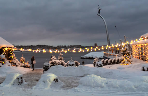 Scenic winter sunset view from the Sørenga neighborhood in Oslo, Norway, showing modern waterfront apartments and pedestrian piers along calm, partially frozen harbor water Warm golden light reflects on the ice and water surface while people walk along the boardwalk creating a peaceful urban atmosphere Sørenga is a contemporary seaside district near the city center known for its canals public sea pool and vibrant residential architecture during a sunny winter evening