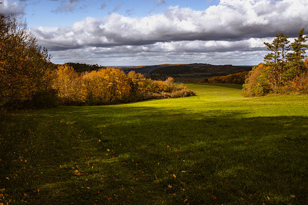 Wide landscape view showing autumn-colored trees, green fields, and a distant village, photographed under partly cloudy skies with natural sunlight.