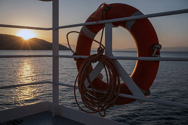 Lifebuoy on ship deck railing against beautiful sea sunset, sun setting behind coastal hill, calm water and warm evening light, concept of cruise travel, safety and peaceful ocean journey