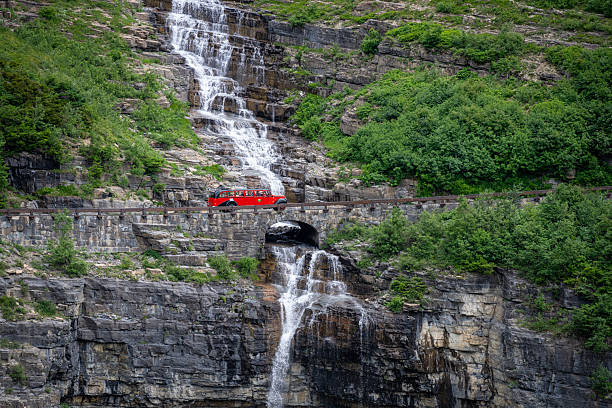 A historic red tour bus travels along Going-to-the-Sun Road past a waterfall and stone bridge in Glacier National Park, Montana, USA, during summer. The scenic mountain roadway is a major tourist route through the park, illustrating sightseeing, transportation, and visitor access in a protected wilderness area.