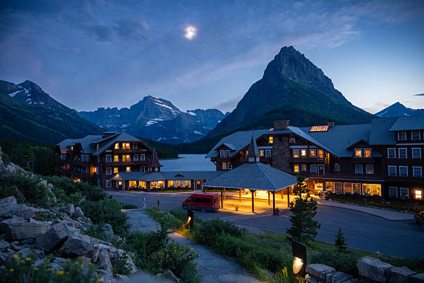 Many Glacier Hotel illuminated at dusk with the moon visible above dramatic alpine peaks in Glacier National Park, Montana, USA. Iconic mountain lodge during blue hour featuring warm lights, rugged Rocky Mountain scenery, and tranquil wilderness atmosphere, ideal for travel, tourism, hospitality, vacation, and destination concepts.