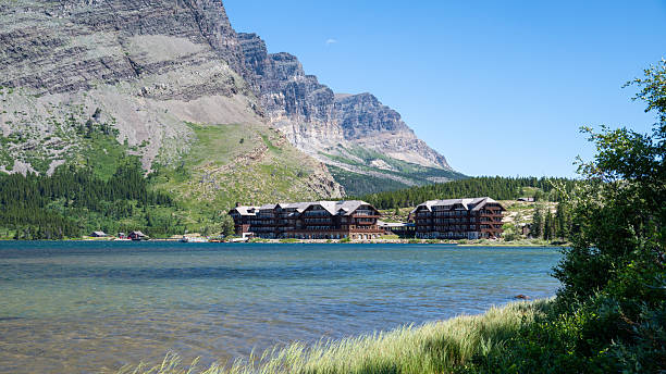 Many Glacier Hotel reflected across an alpine lake beneath dramatic mountain cliffs in Glacier National Park. Scenic summer landscape with clear water, green shoreline vegetation, evergreen forest, and rugged Rocky Mountain scenery, ideal for travel, tourism, hospitality, vacation, and destination concepts.