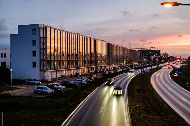 Amersfoort, The Netherlands - January 16th, 2026: Modern building alongside a busy urban road at dusk, captured from an elevated viewpoint. Passing cars create light trails due to long exposure, while warm interior lights reflect in the glass facade. The image conveys themes of urban life, mobility, architecture, and evening traffic in a modern city environment.