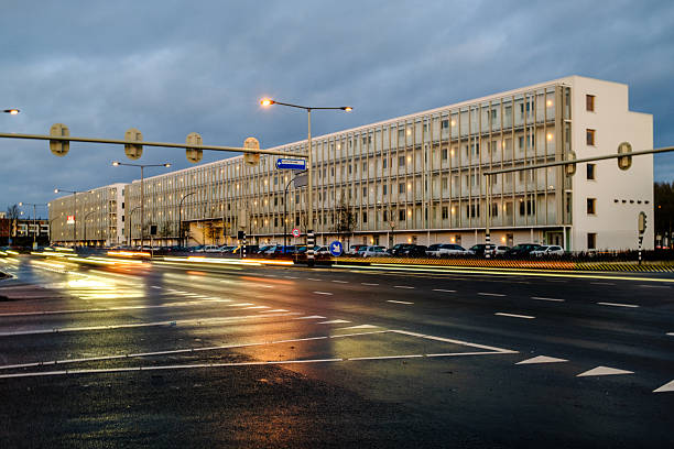 Amersfoort, The Netherlands - January 16th, 2026: Modern building alongside a busy urban road at dusk, captured from an elevated viewpoint. Passing cars create light trails due to long exposure, while warm interior lights reflect in the glass facade. The image conveys themes of urban life, mobility, architecture, and evening traffic in a modern city environment.