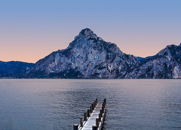 Scenic winter view of a snow-covered wooden pier leading towards the majestic Traunstein mountain on Lake Traunsee during a peaceful sunrise in Traunkirchen, Austria