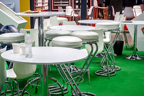 This image captures a contemporary cafe or lounge setting, featuring an array of sleek white round tables and comfortable white cushioned bar stools. The vibrant green carpet provides a striking contrast, enhancing the modern aesthetic of the space. With chrome accents on the table bases and stool legs, the design emphasizes clean lines and a minimalist feel. This versatile setup could represent a coffee break area at a conference, an exhibition booth, an airport lounge, or a trendy restaurant interior, offering a comfortable and stylish environment for casual gatherings or refreshments.