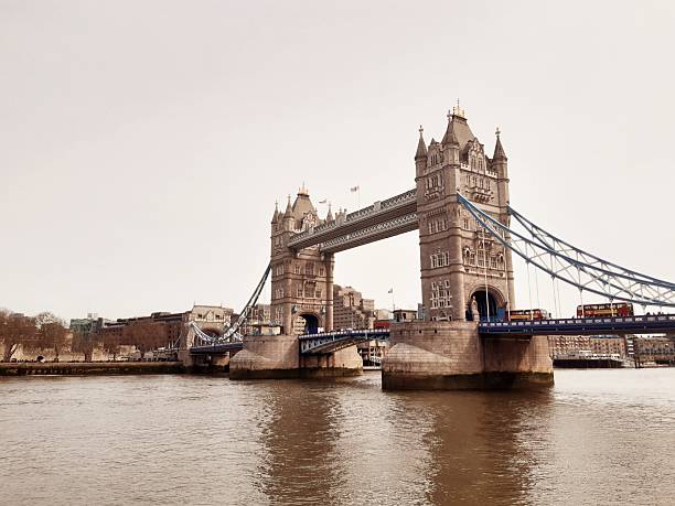 A wide landscape view of Tower Bridge, showing the two neo-Gothic towers and the blue suspension cables. Red double-decker buses are visible crossing the bridge over the River Thames under an overcast sky