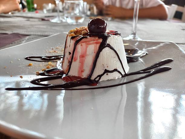 Dessert individuel dressé sur assiette blanche : un cylindre de mousse blanche garni de miettes croustillantes, nappé de coulis de fruits rouges et de sauce chocolat, surmonté d’une cerise confite, avec décor de chocolat autour.