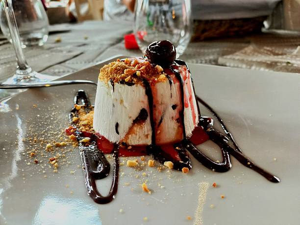 Dessert individuel dressé sur assiette blanche : un cylindre de mousse blanche garni de miettes croustillantes, nappé de coulis de fruits rouges et de sauce chocolat, surmonté d’une cerise confite, avec décor de chocolat autour.