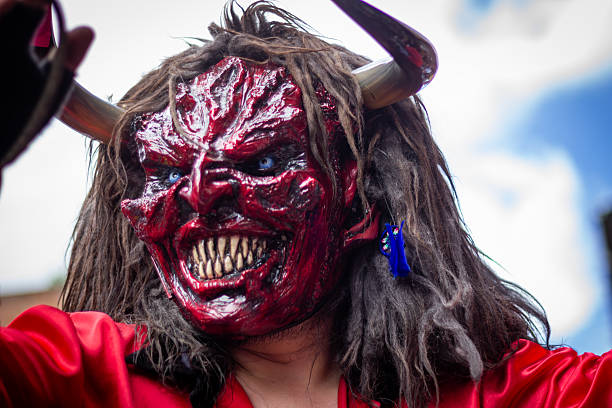 Scary red demon mask with horns, sharp teeth and glowing blue eyes worn at an outdoor festival, traditional folklore costume performance evoking horror and celebration