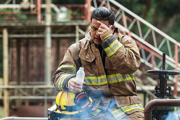 Asian firefighter wearing protective uniform drinking water during a break at an industrial site. Concept of hard work, hydration, safety, emergency service, industrial labor, and recovery after intense duty.