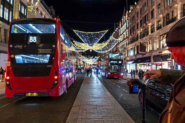 London, United Kingdom: A nighttime urban street scene on Brompton Road in the Knightsbridge area captures red double-decker buses, including route 88, traveling along a wet pavement illuminated by strings of colorful Christmas lights overhead. Pedestrians, including a middle-aged man with a beard wearing a brown coat and hat gesturing with his hand, walk on the sidewalk amidst the holiday decorations. Luxury storefronts such as Harrods are visible on both sides of the street, with additional buses and cars in the background. The atmosphere reflects winter evening activity in a bustling shopping district during the festive season.