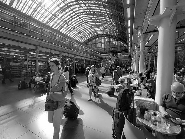 London, UK:A diverse group of travellers on the Elizabeth underground tube station, including men and women of various ages and ethnicities, navigate a spacious indoor tube station. Several individuals carry backpacks and walk along the polished floor or ascend and descend multiple escalators. The setting features contemporary architecture with extensive glass panelling, metallic escalator structures, and large digital display screens showing information. Red barriers line some areas, and the open atrium design creates a sense of height and modernity. The scene captures everyday commuter and passenger movement in a busy transit hub, with neutral expressions focused on travel. No specific time of day or season is evident, emphasizing the functional and efficient environment of air travel infrastructure.