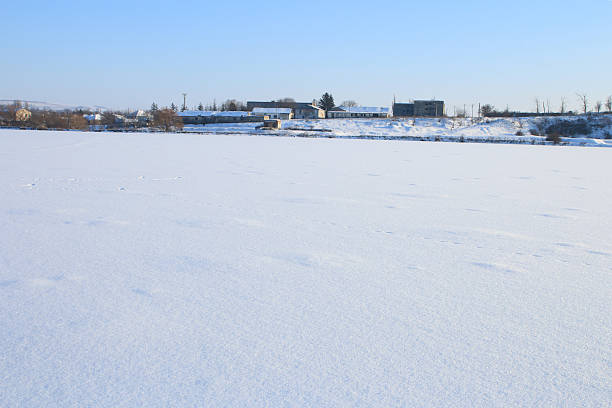 A snowy field with buildings in the background. Sky. Outdoor. Snow. Nature. Shore