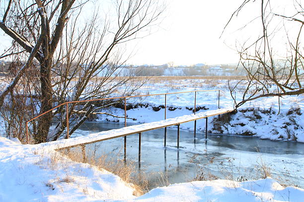 A bridge over a river with snow on the banks. Tree. Snow. Outdoor. Nature. Pond