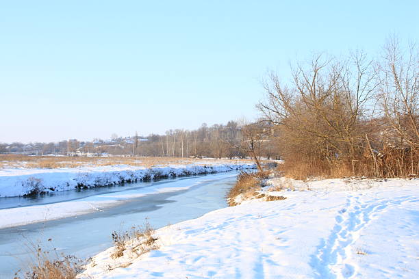 A snowy field with trees and a road with snow on it. Outdoor. Snow. Sky. Nature. Shore