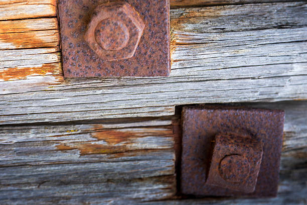 Under the pier, drift wood, wooden textures and rusty nails. Weathered wooden beams with visible grain and texture support a pier. Rusty metal bolts and nails secure the structure, displaying a combination of weathering and corrosion. The scene suggests exposure to marine elements, with the beach in the background. The aged wood and rust create a rustic, historical appearance, evoking the harsh conditions typical of coastal environments.