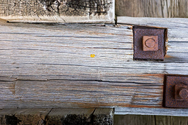 Under the pier, drift wood, wooden textures and rusty nails. Weathered wooden beams with visible grain and texture support a pier. Rusty metal bolts and nails secure the structure, displaying a combination of weathering and corrosion. The scene suggests exposure to marine elements, with the beach in the background. The aged wood and rust create a rustic, historical appearance, evoking the harsh conditions typical of coastal environments.